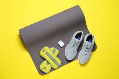 Photo of Exercise mat, weights, wireless earphones and shoes on yellow background, top view