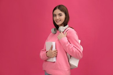 Teenage student with book, headphones and backpack on pink background