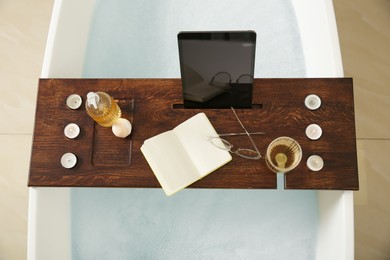 Wooden bath tray with candles, book and tablet on tub indoors, top view
