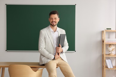 Happy teacher with stationery at desk in classroom