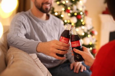 MYKOLAIV, UKRAINE - JANUARY 27, 2021: Young couple holding bottles of Coca-Cola in room decorated for Christmas, closeup