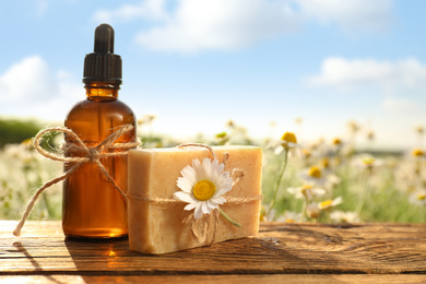 Bottle of chamomile essential oil and soap bar on wooden table in field