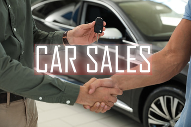 Young salesman shaking hands with customer and giving car key in modern auto dealership, closeup