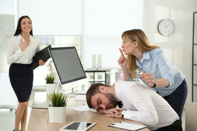 Young woman sticking paper fish to colleague's back while he sleeping in office. Funny joke