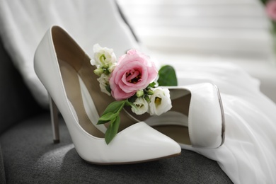 Pair of white high heel shoes, flowers and wedding dress on chair indoors, closeup