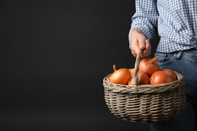Woman holding wicker basket with raw yellow onion bulbs on black background, closeup. Space for text