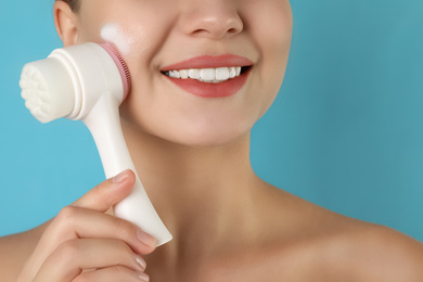 Young woman washing face with brush and cleansing foam on light blue background, closeup. Cosmetic products