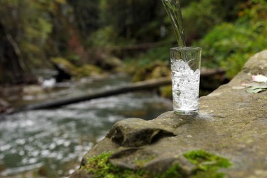 Fresh water pouring into glass on stone near stream in forest. Space for text