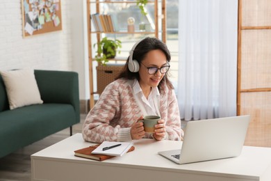 Woman with modern laptop and headphones drinking tea while learning at table indoors