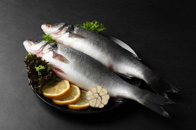 Photo of Plate with fresh raw sea bass fish and ingredients on black table