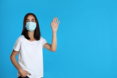Photo of Young woman in protective mask showing hello gesture on light blue background, space for text. Keeping social distance during coronavirus pandemic