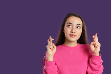 Woman with crossed fingers on purple background, space for text. Superstition concept