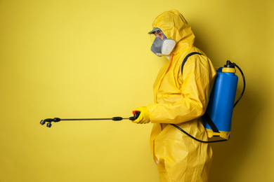 Man wearing protective suit with insecticide sprayer on yellow background. Pest control
