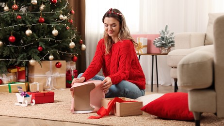Beautiful young woman wrapping gift near Christmas tree at home