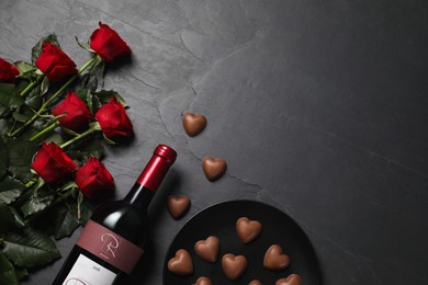 Photo of Bottle of red wine, beautiful roses and heart shaped chocolate candies on black table, flat lay. Space for text