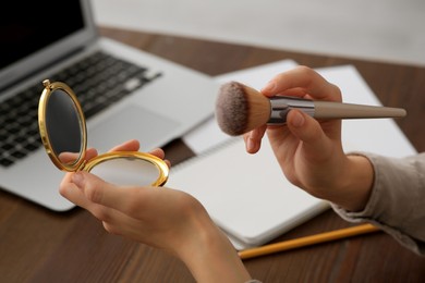 Woman with cosmetic pocket mirror and makeup brush at wooden table indoors, closeup