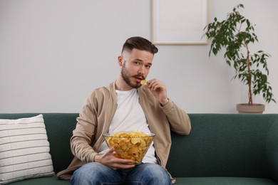 Handsome young man eating tasty potato chips on sofa at home
