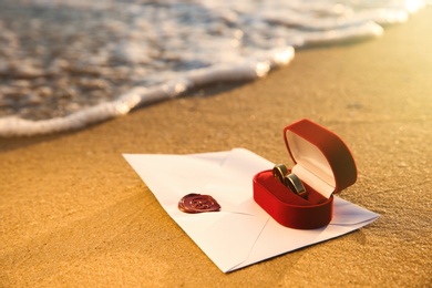 Red box with gold wedding rings and envelope on sandy beach