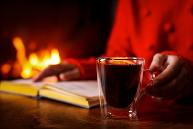 Woman with tasty mulled wine reading book near fireplace at home, closeup