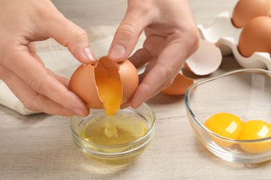 Woman separating egg yolk from white over glass bowl at wooden table, closeup