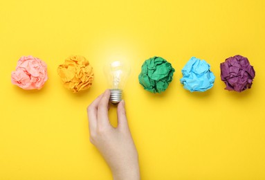 Woman holding lightbulb among colorful paper balls on yellow background, top view. Idea concept