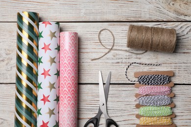 Different colorful wrapping paper rolls, scissors and threads on white wooden table, flat lay