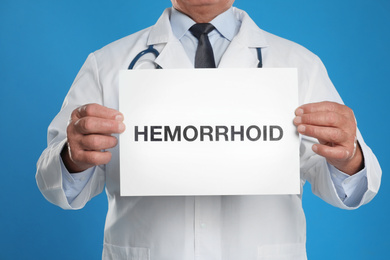 Doctor holding sign with word HEMORRHOID on blue background, closeup