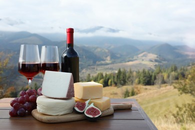 Different types of delicious cheeses, snacks and wine on wooden table against mountain landscape. Space for text