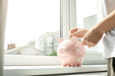 Woman putting money into piggy bank at window sill indoors, closeup. Space for text