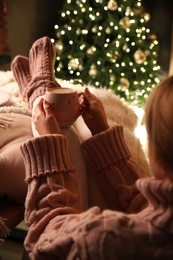 Woman holding cup of delicious hot drink near Christmas tree indoors, closeup