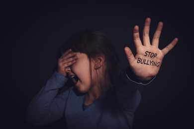 Crying little girl showing palm with message STOP BULLYING near black wall, focus on hand