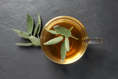 Cup of aromatic sage tea and fresh leaves on black table, flat lay