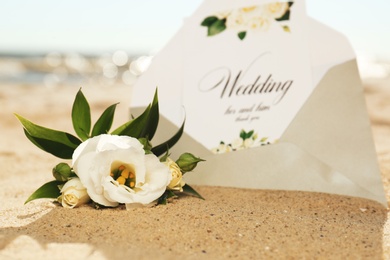 Envelope with wedding invitation and beautiful flower on sandy beach, closeup