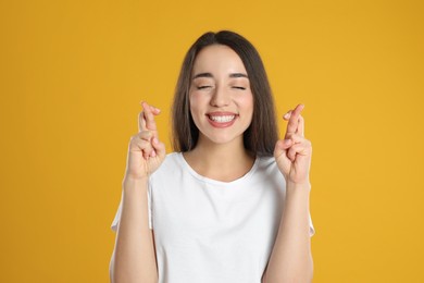 Excited young woman holding fingers crossed on yellow background. Superstition for good luck