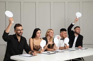 Panel of judges with different emotions holding blank score signs at table against light wall. Space for text