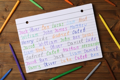 Photo of Notebook with different baby names and colorful markers on wooden table, flat lay