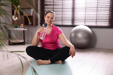 Overweight woman with bottle of water on yoga mat at home