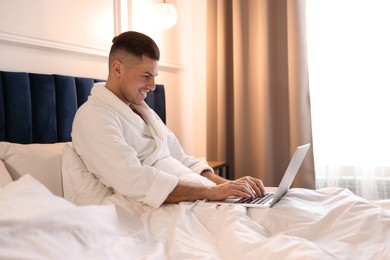 Handsome man wearing bathrobe with laptop on bed in hotel room
