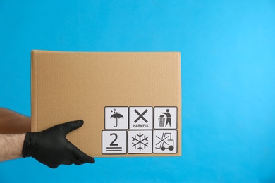 Courier holding cardboard box with different packaging symbols on blue background, closeup. Parcel delivery