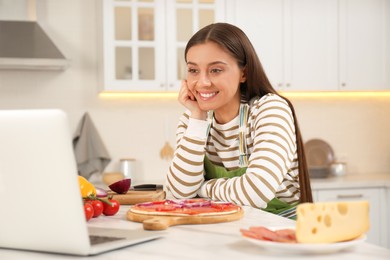 Happy woman watching online cooking course via laptop in kitchen