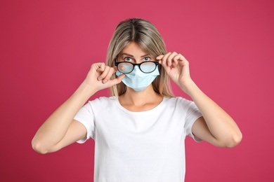 Woman wiping foggy glasses caused by wearing disposable mask on pink background. Protective measure during coronavirus pandemic
