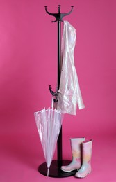 Stylish rack with umbrella, raincoat and rubber boots on pink background