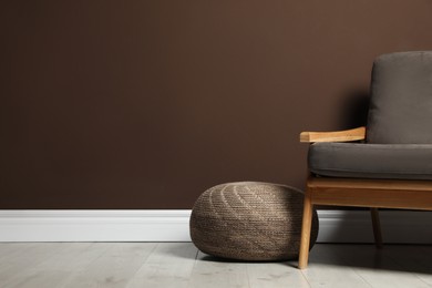 Stylish knitted pouf and comfortable armchair near brown wall indoors, space for text. Interior design