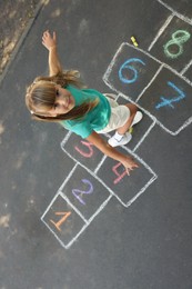Photo of Little girl playing hopscotch drawn with chalk on asphalt outdoors, above view. Happy childhood
