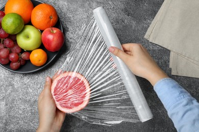 Woman putting plastic food wrap over half of grapefruit at grey table, top view
