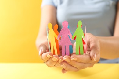 Photo of Woman holding paper human figures at yellow table, closeup. Diversity and inclusion concept