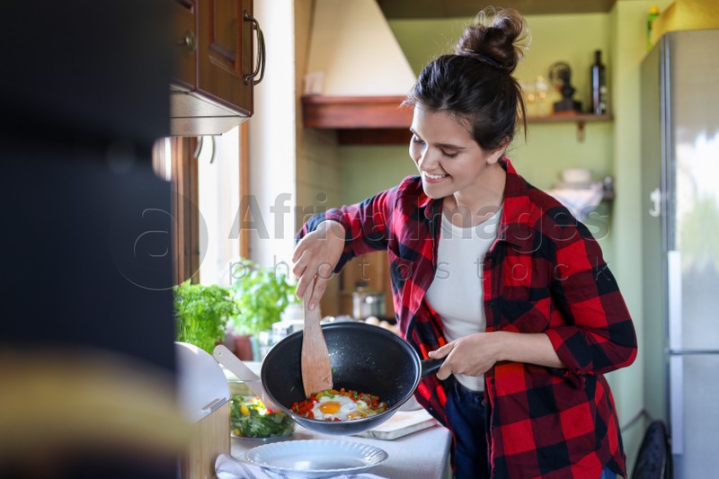 Photo of Young woman putting freshly fried eggs and vegetables onto plate in kitchen