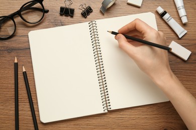 Woman with pencil and blank sketchbook at wooden table, top view