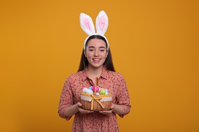 Photo of Happy woman in bunny ears headband holding wicker basket of painted Easter eggs on orange background