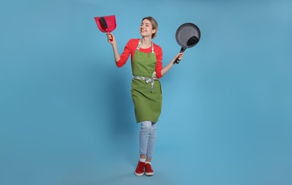 Young housewife with dustpan, brush, frying pan and spatula on light blue background